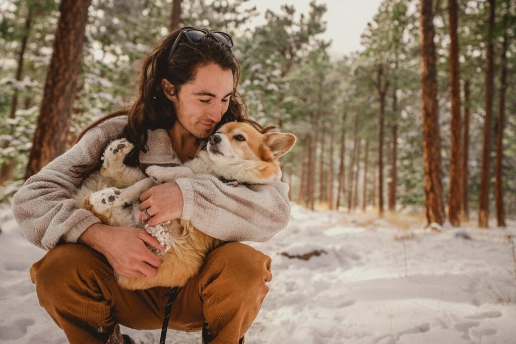 Man with brown hair cradling his light brown haired corgi in the snowy woods.