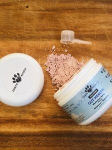image of an open jar of earth buddy gut health with colostrum powder on wood table with a scoop and lid laying next to the jar. Colostrum for dogs is a great natural remedy for skin issues.