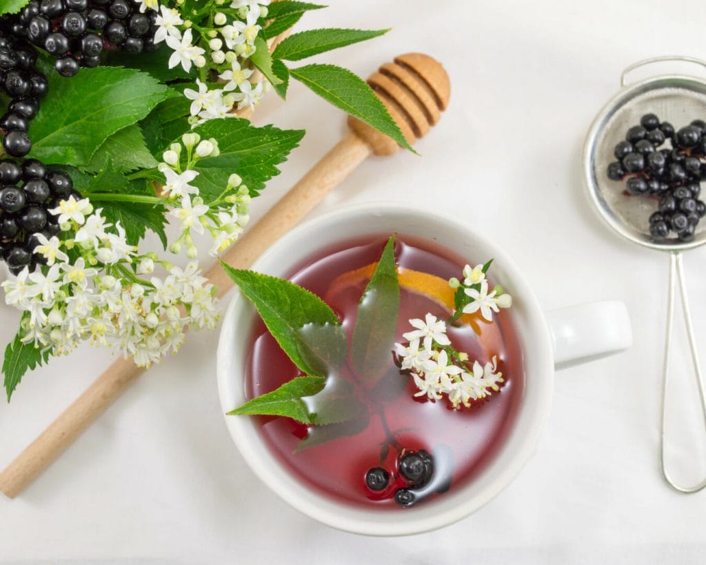 image of elderberry flower, berries, and elderberry extract in a bowl with kitchen utensils. Elderberries are high in vitamin C and quercetin.