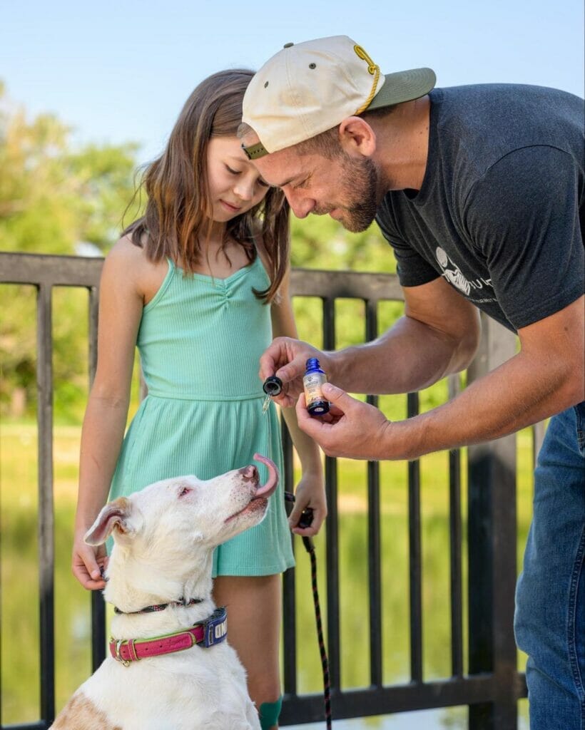 Earth Buddy founder, Sean Zyer and his daughter giving Cellular Support 500mg CBG Extract to white dog with brown spots. CBG for dogs helps with IBS and seizures.