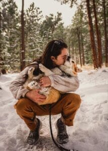 Owner holding a tan & white Corgi dog in the woods of the Rocky Mountains. Try using organic CBD oil for dogs when camping to help with stress. 
