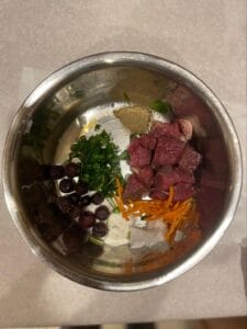 Dog food bowl with raw ingredients like raw meat, blueberries, carrots, greens, and pet supplements to support a holistic dog diet. 