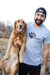 Man in an Earth Buddy T-shirt holding a happy golden retriever under his arm. Earth Buddy is the best cbd oil for dogs.