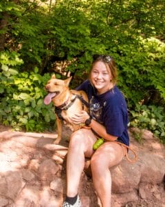 Woman with a tan, mixed breed dog with harness in the woods. Read this blog to learn how cbd oil helps stressed dogs.