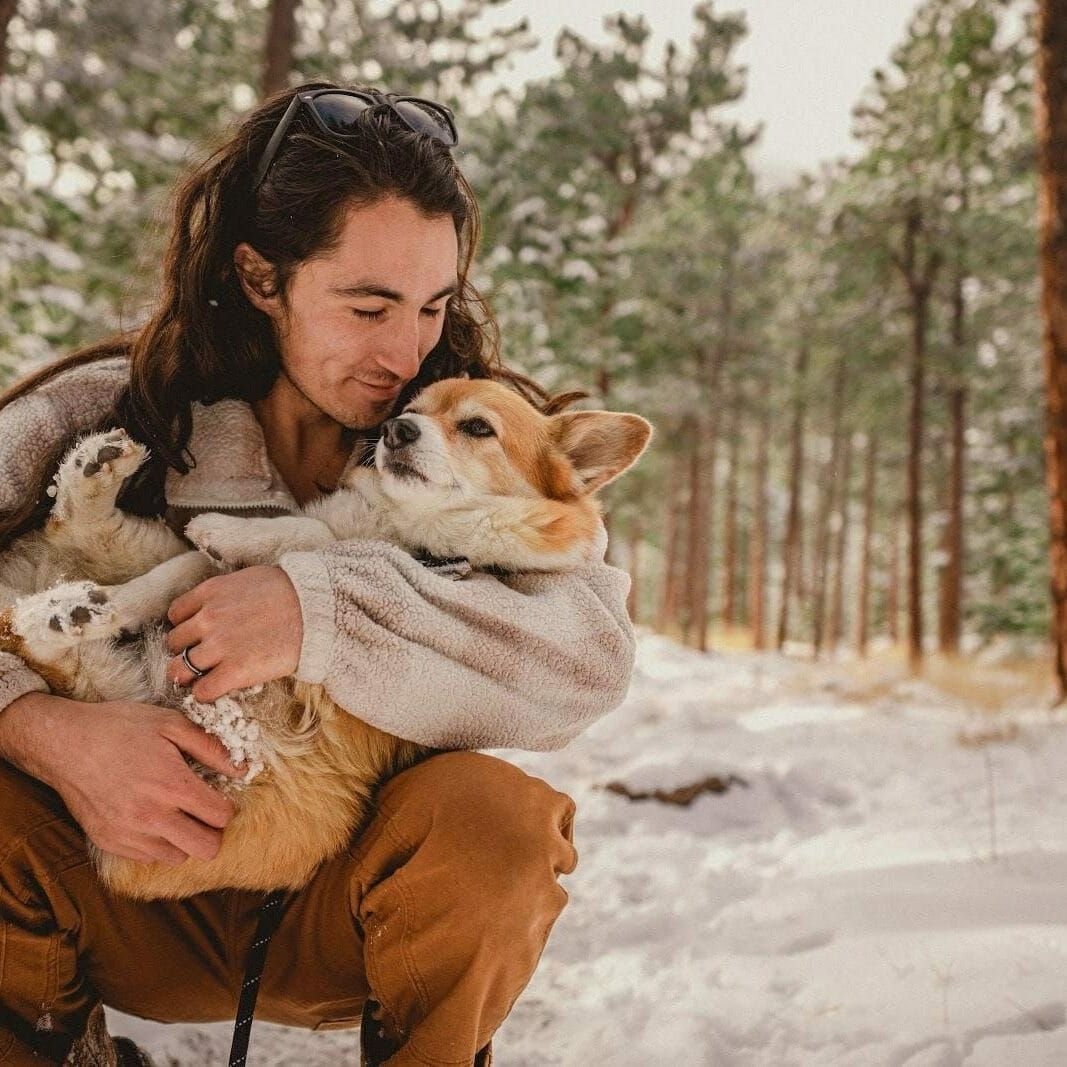 Man with brown hair cradling his light brown haired corgi in the snowy woods.