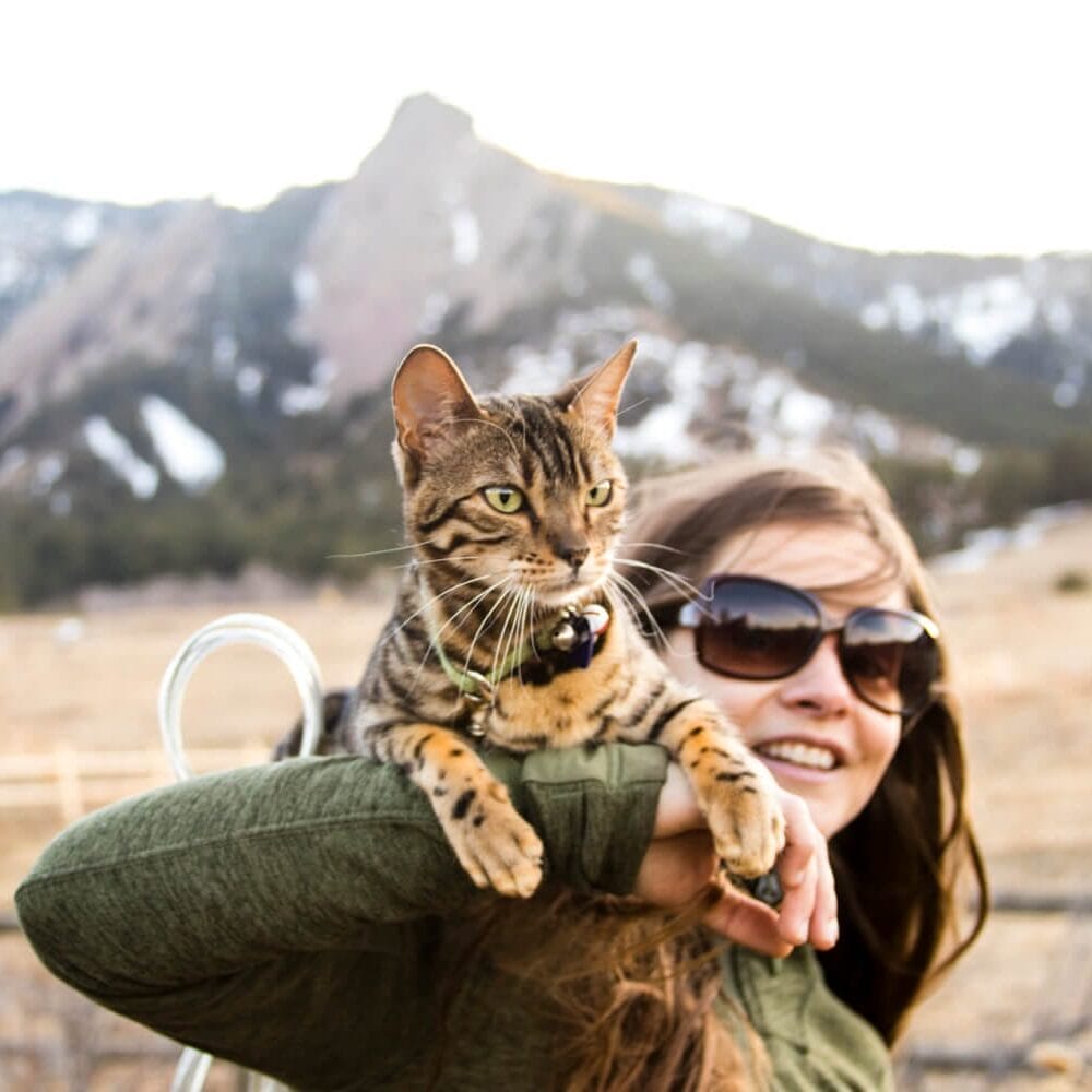 Tan Bengal Cat on the shoulders of a woman outdoors in the rocky mountains. If your cat has a dull coat, read this blog to learn about cat supplements for a healthy coat.