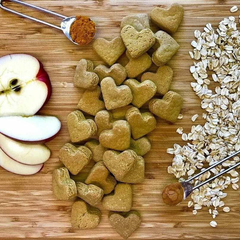 light brown cutting board with whole food ingredients Earth Buddy uses in their CBD treats for dogs like apples, cinnamon, oats, and finished cbd dog treats.